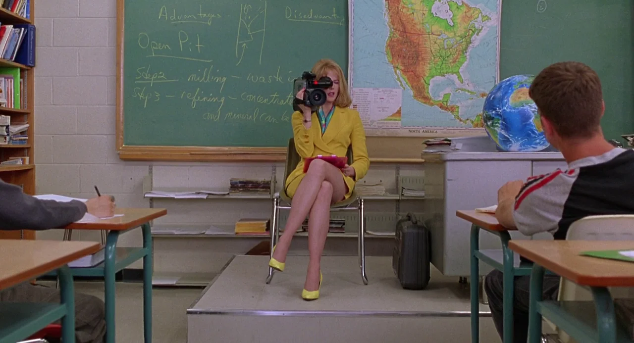 To Die For (1995) movie still: medium — A school classroom features a green chalkboard with notes and a world map. A woman in a yellow suit sits on…