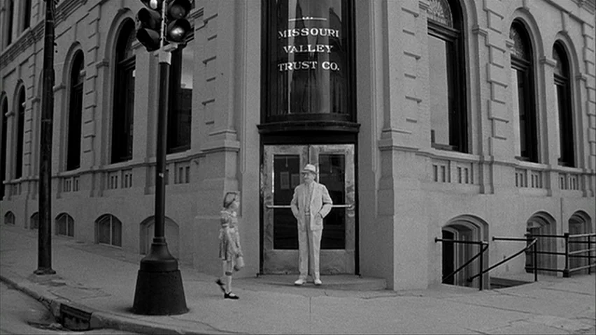 Paper Moon (1971) movie still: wide — The corner of a stone bank building features tall windows and a glass door. An elderly man in a suit stands at…