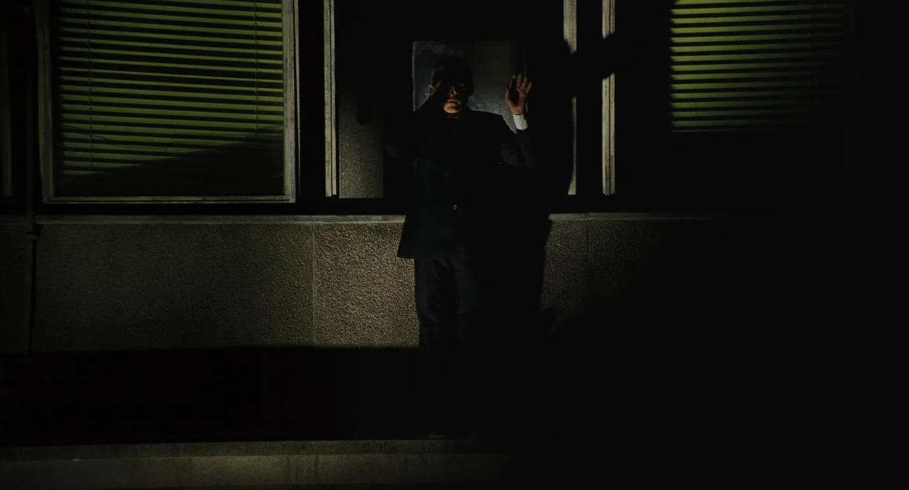 The Double (2013) movie still: fullbody — An exterior concrete building wall features two dark-tinted windows with closed green blinds. A man in a…