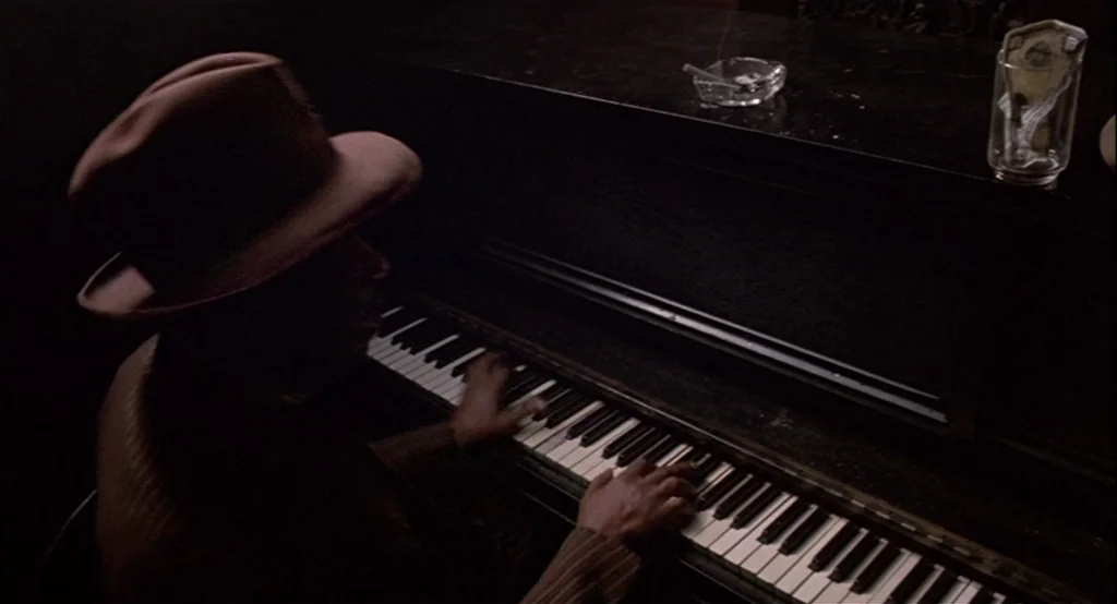 The Cotton Club (1984) movie still: medium — A dimly lit room features a black piano with a glass ashtray and jar on top. A man wearing a fedora hat…