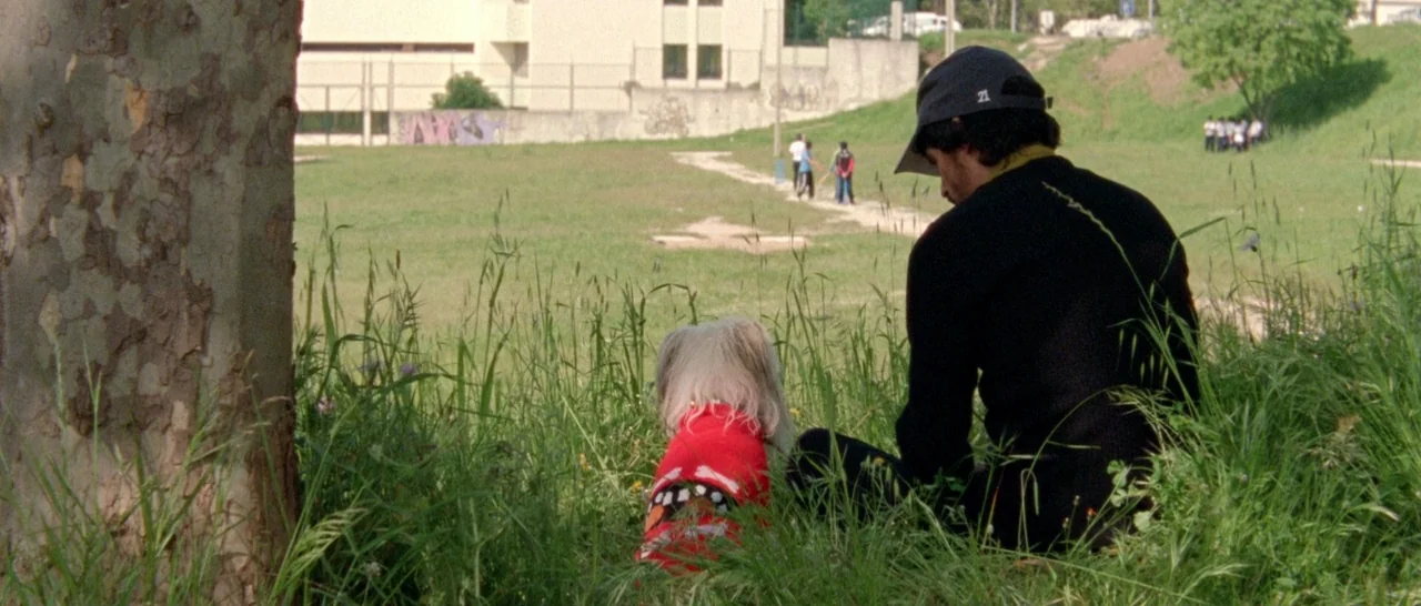 Arabian Nights: Volume 2 The Desolate One (2015) movie still: medium — An open grassy area sits adjacent to a concrete building with a low wall. A…