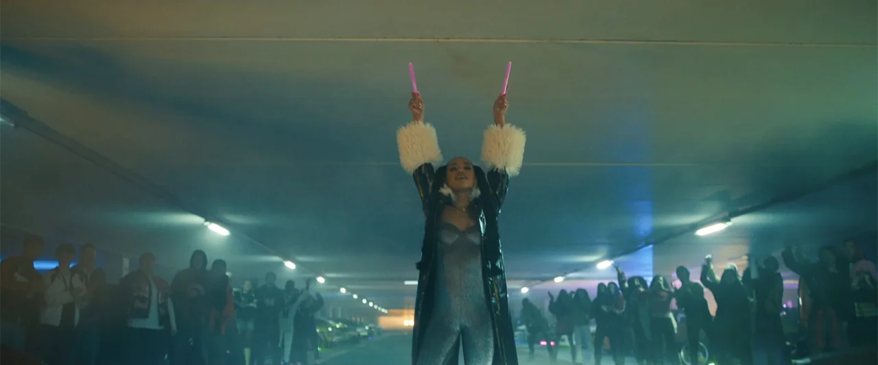 My Fault: London (2025) movie still: fullbody — A concrete parking garage is populated by a crowd gathered around a central figure. A young woman in…