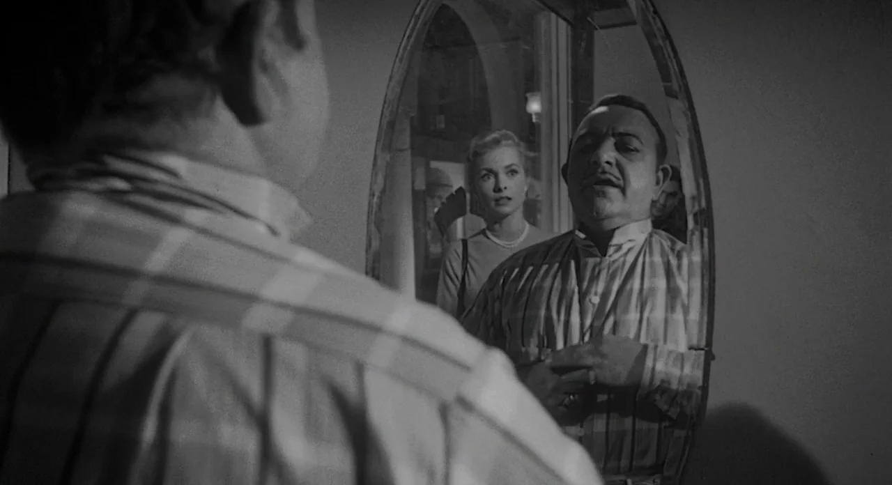 Touch of Evil (1958) movie still: medium — A man looks into a tall oval mirror while a woman stands behind him. The room is dimly lit, reflecting…