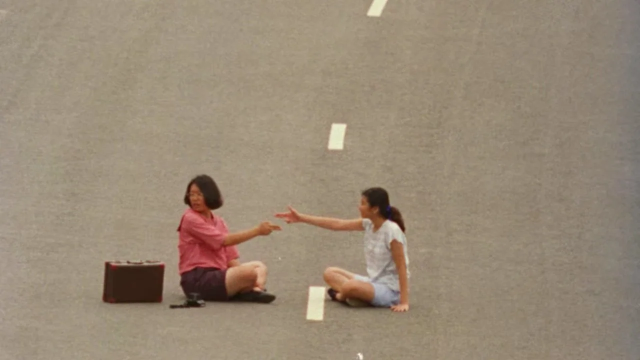 Shirkers (2018) movie still: wide — An empty stretch of paved asphalt road with dashed white lane markings. Two women sit facing each other on the…