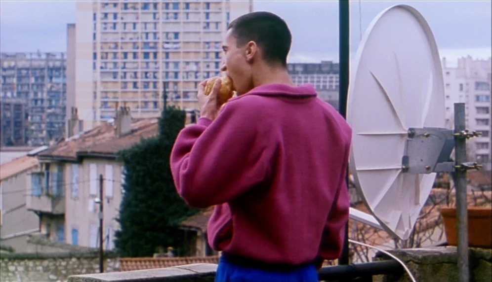 Nenette et Boni (1996) movie still: medium — A residential balcony overlooks a dense urban landscape of apartment blocks and houses. A young man…