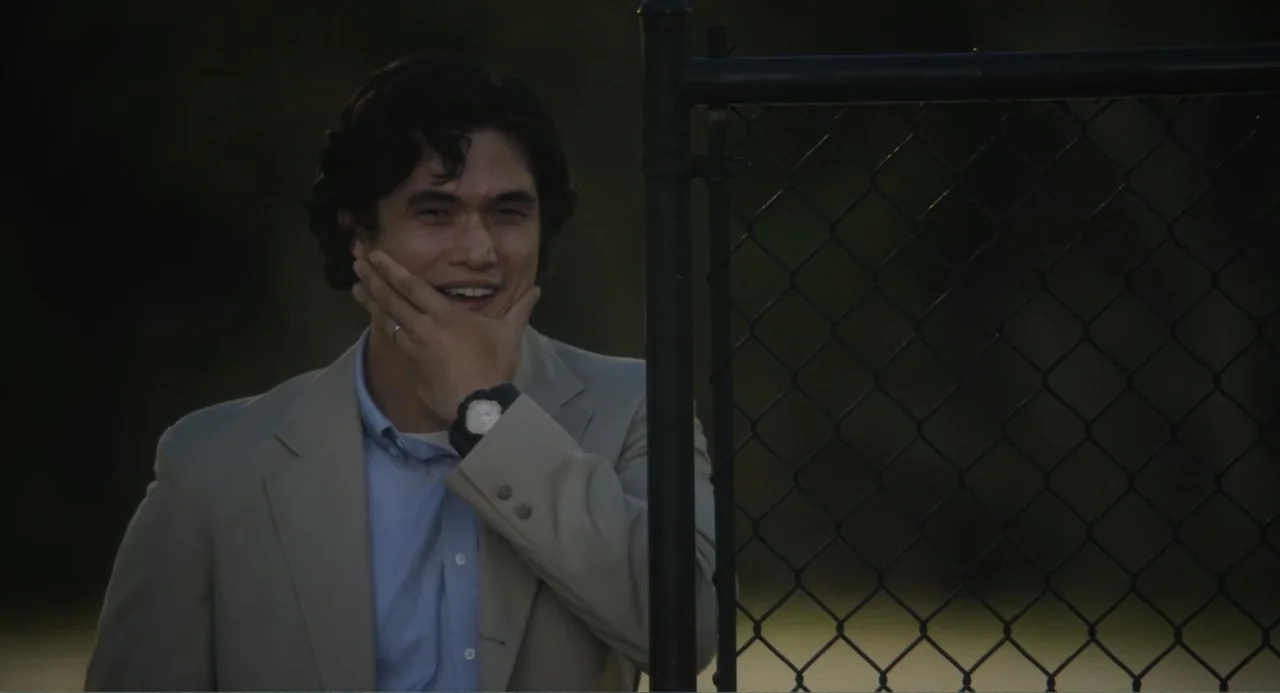 May December (2023) movie still: medium — A man stands outdoors next to a black chain-link fence during late afternoon. He is touching his jaw with…