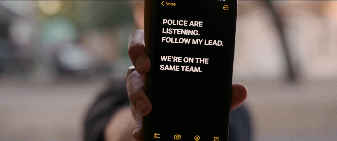 Hit Man (2023) movie still: closeup — A person holds a smartphone screen showing a notes application in an outdoor setting. The text on the screen…
