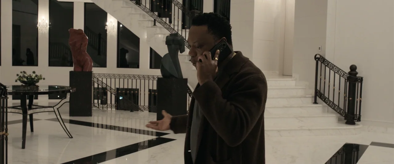 Play Dirty (2025) movie still: medium — A spacious foyer with polished white marble floors, black tile inlays, and curved staircases. A man in a…