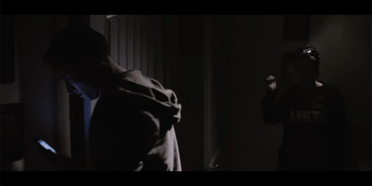 La coda del diavolo (2024) movie still: medium — A dark residential hallway contains two figures standing in low light. One man checks his glowing…