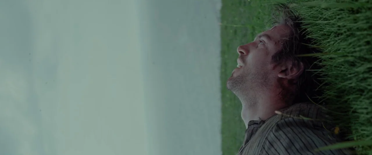 Apostle (2018) movie still: closeup — An open field with tall green grass stretches under an overcast grey sky. A man lies horizontally in the grass…