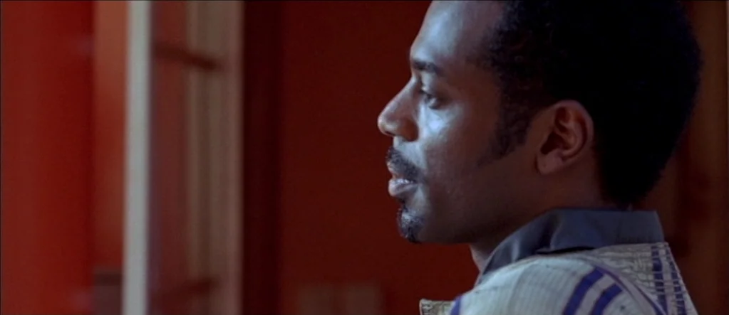 White Material (2009) movie still: closeup — A man stands in an interior space with warm, dark red walls. He looks towards the left of the frame in…