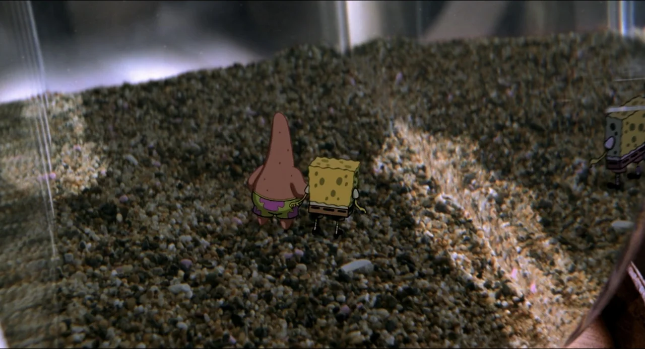The SpongeBob SquarePants Movie (2004) movie still: wide — A glass container holds a layer of small grey and brown aquarium gravel pebbles. Two…
