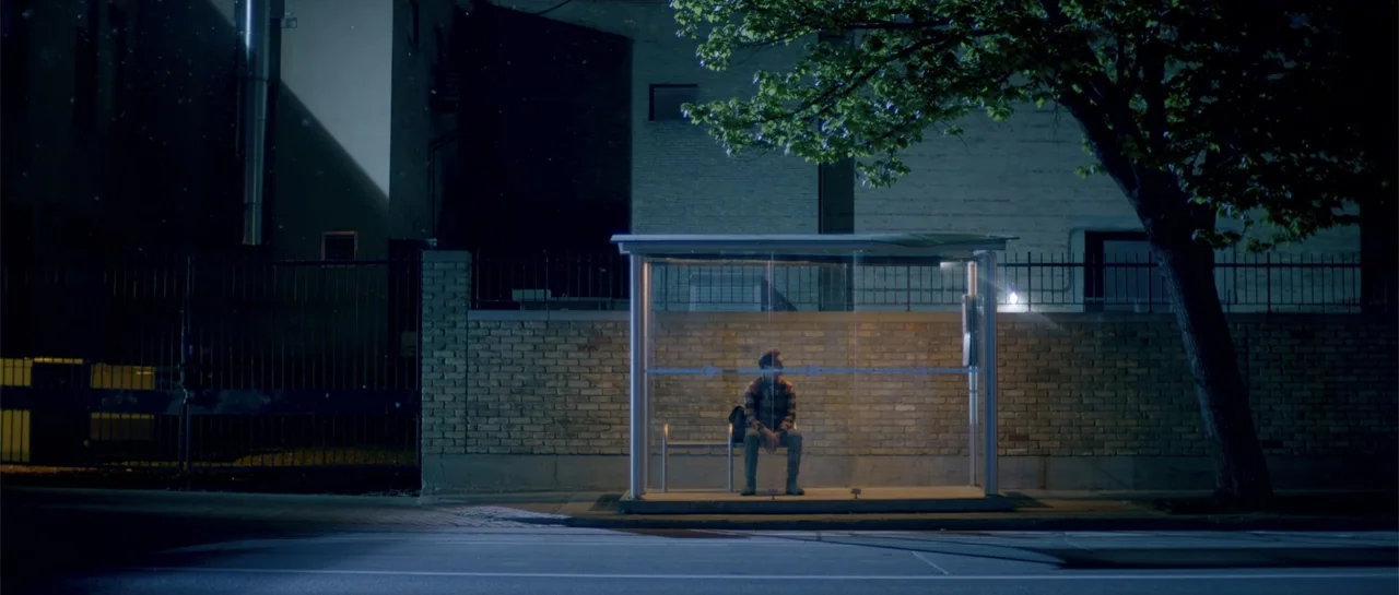 Elevator Game (2023) movie still: wide — A modern glass bus shelter stands on a curb at night against a brick wall. A man sits alone on the metal…