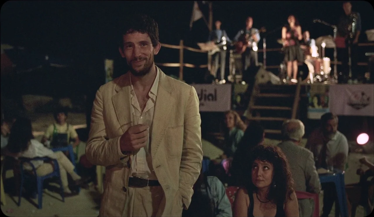 La Chimera (2023) movie still: medium — An outdoor nighttime gathering features a live band performing on a small raised stage. A man in a beige…