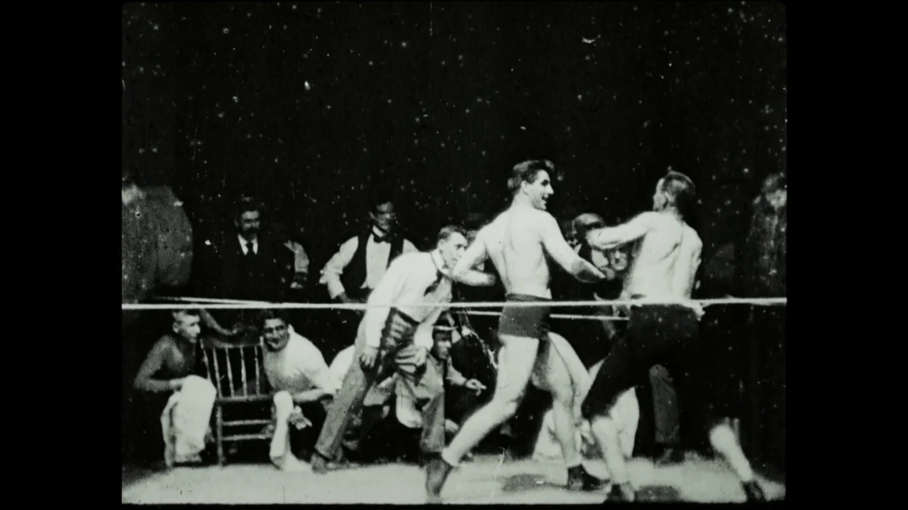 Hugo (2011) movie still: fullbody — A dark, dimly lit indoor space houses a roped-off boxing fighting area. Two men in dark trunks exchange blows…