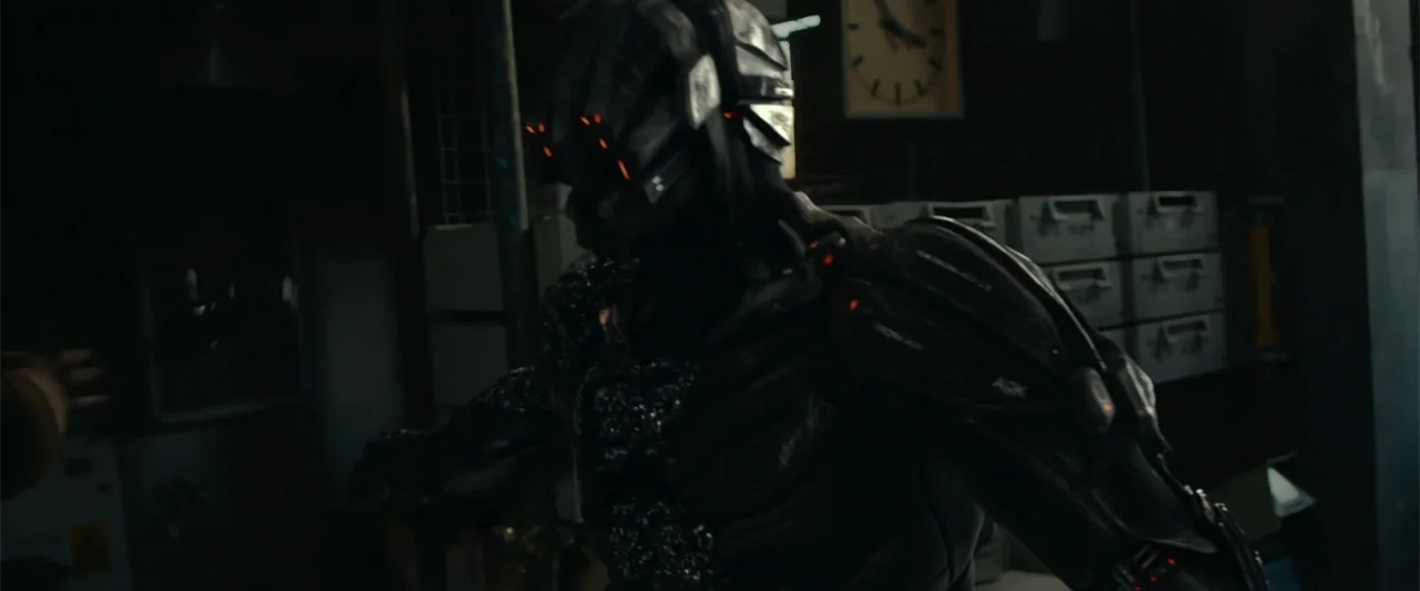 The Condemned 2 (2015) movie still: medium — A dark, cluttered room contains storage cabinets and an analog wall clock. A futuristic armored figure…