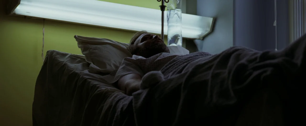 Syriana (2005) movie still: medium — A hospital bed with a white pillow and sheet sits against a yellowish wall. A bearded man in a hospital gown…
