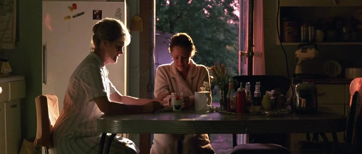 At Close Range (1986) movie still: medium — A rustic kitchen features a metal-rimmed table and an open door to evening light. Two women sit at the…