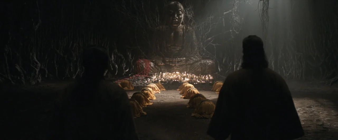 47 Ronin (2013) movie still: wide — A dimly lit cavern contains a massive weathered stone Buddha statue surrounded by dark roots. Multiple figures…