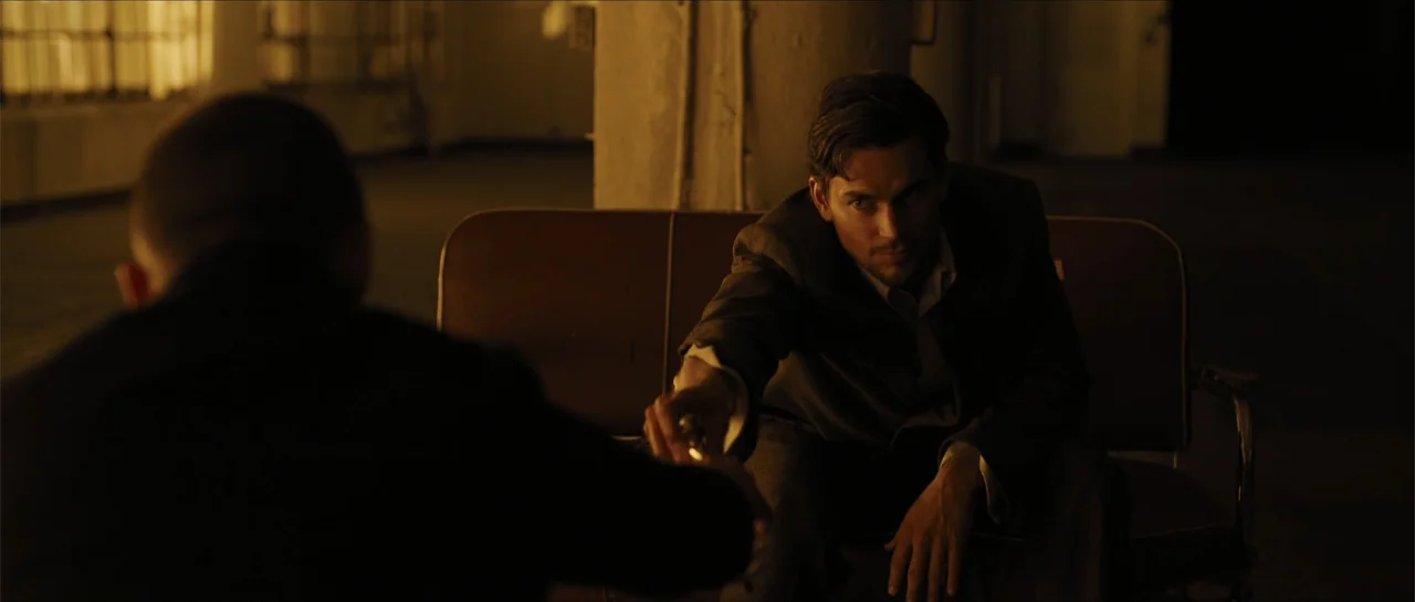 In Time (2011) movie still: medium — A dimly lit room with an indistinct backdrop and a vinyl bench seat. A man in a suit reaches forward to…