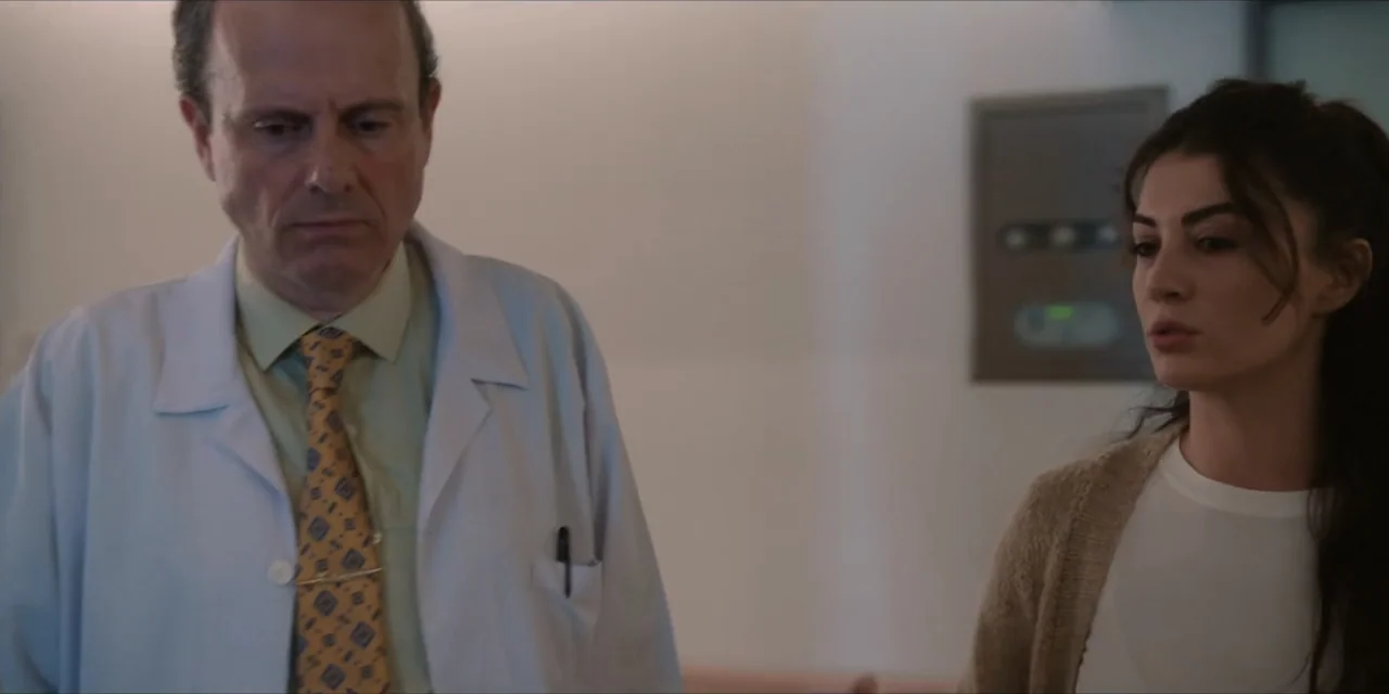 Kill Me If You Dare (2019) movie still: medium — A hospital hallway features plain light-colored walls and a wall-mounted electronic display panel.…