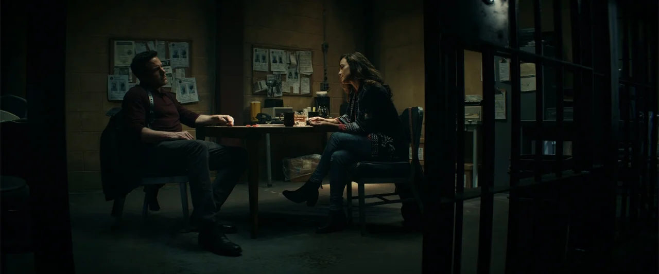 Hypnotic (2023) movie still: medium — An interrogation room features cinder block walls decorated with pinned paperwork and metal bars. A man and a…