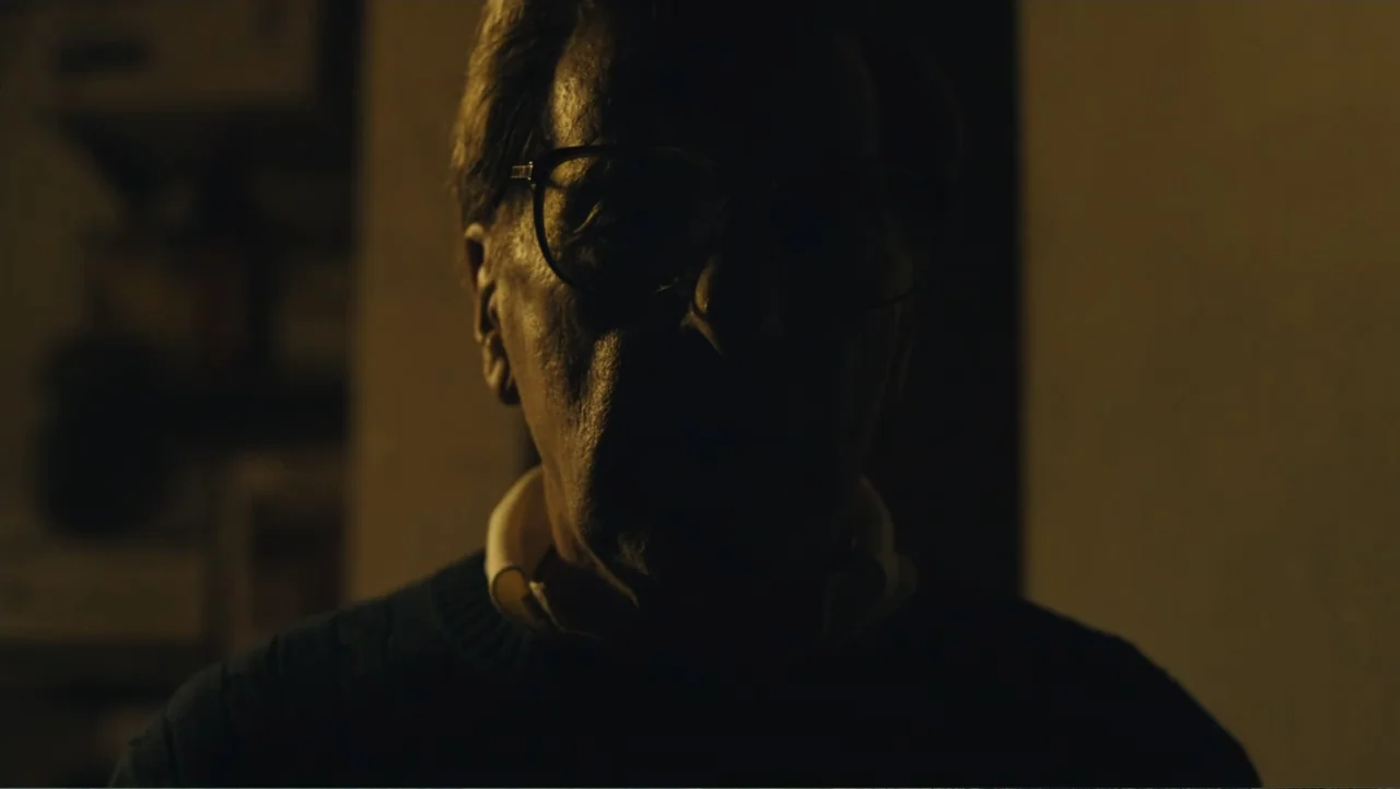 Paterno (2018) movie still: closeup — An interior dimly lit room features a mature man standing before the camera lens. He maintains a steady gaze…