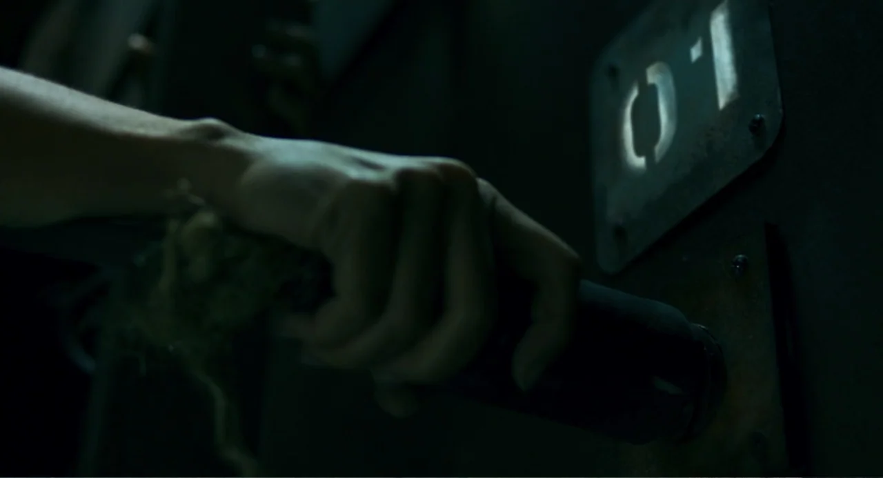 The Lift (2021) movie still: closeup — A dark, metallic surface features a wall-mounted handle and a small numeric identification plate. A human…