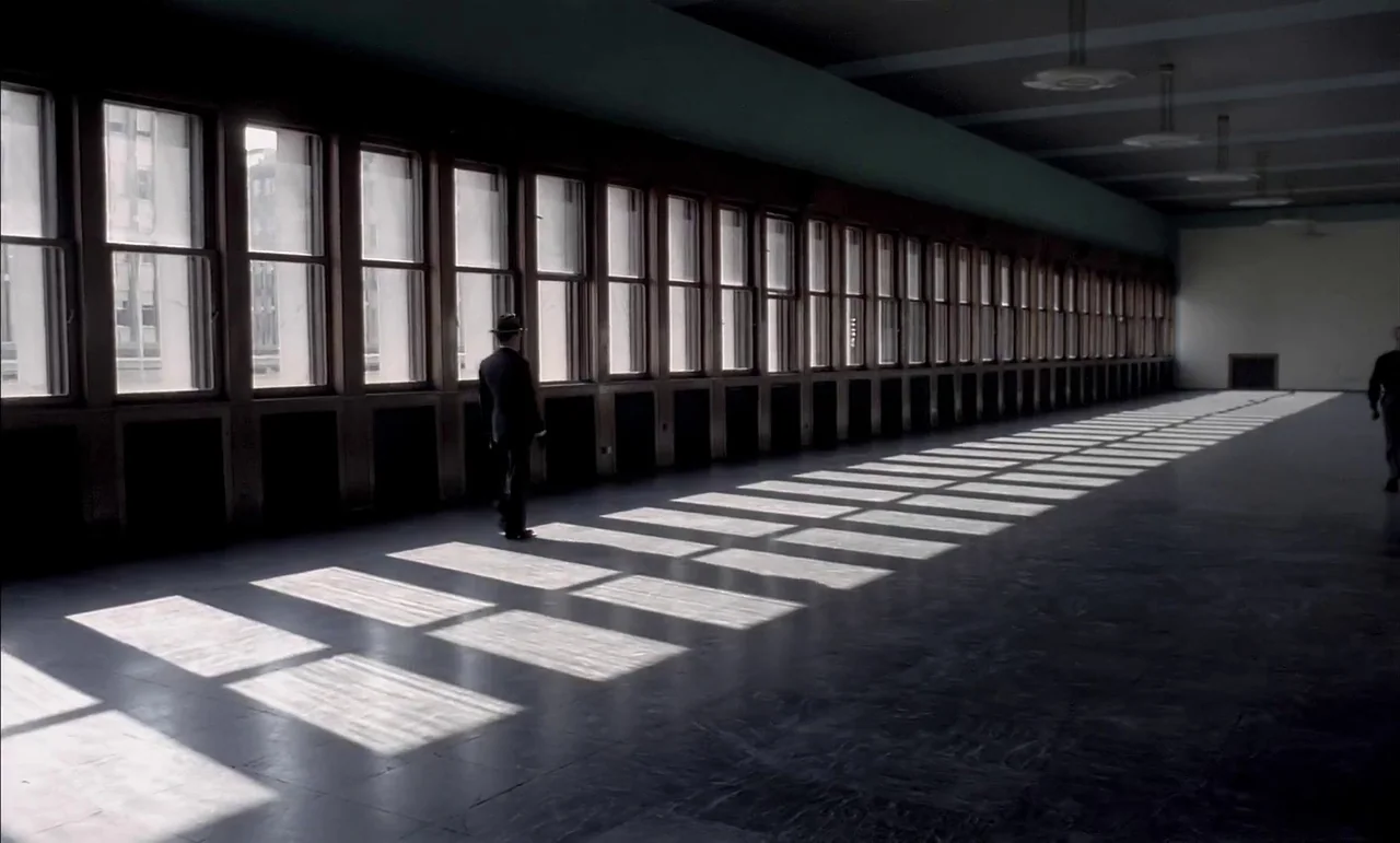 The Conformist (1970) movie still: wide — A long, minimalist hallway features a bank of tall, rectangular windows along one side. A man wearing a…