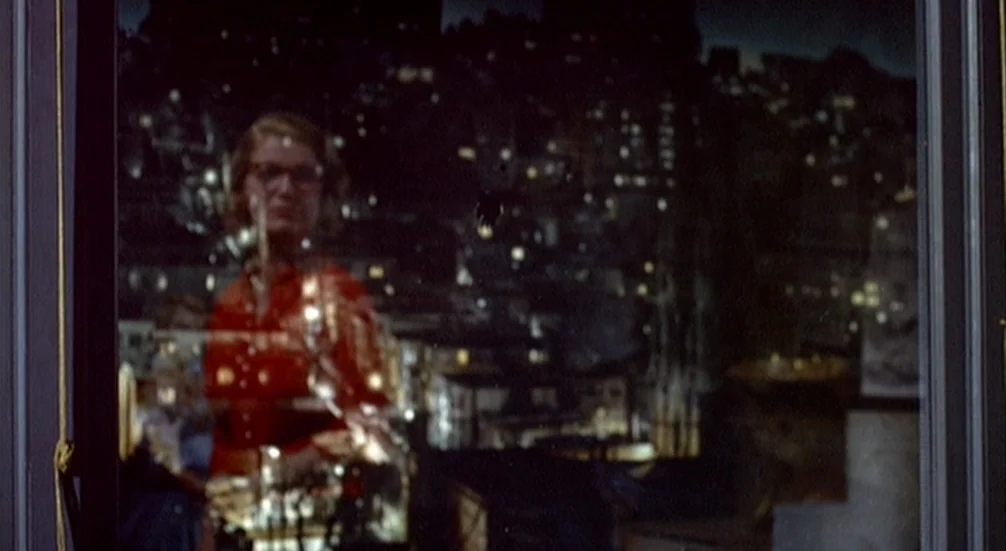Vertigo (1958) movie still: medium — A woman stands behind a window looking out at a dark city landscape at night. The reflection of her face and…