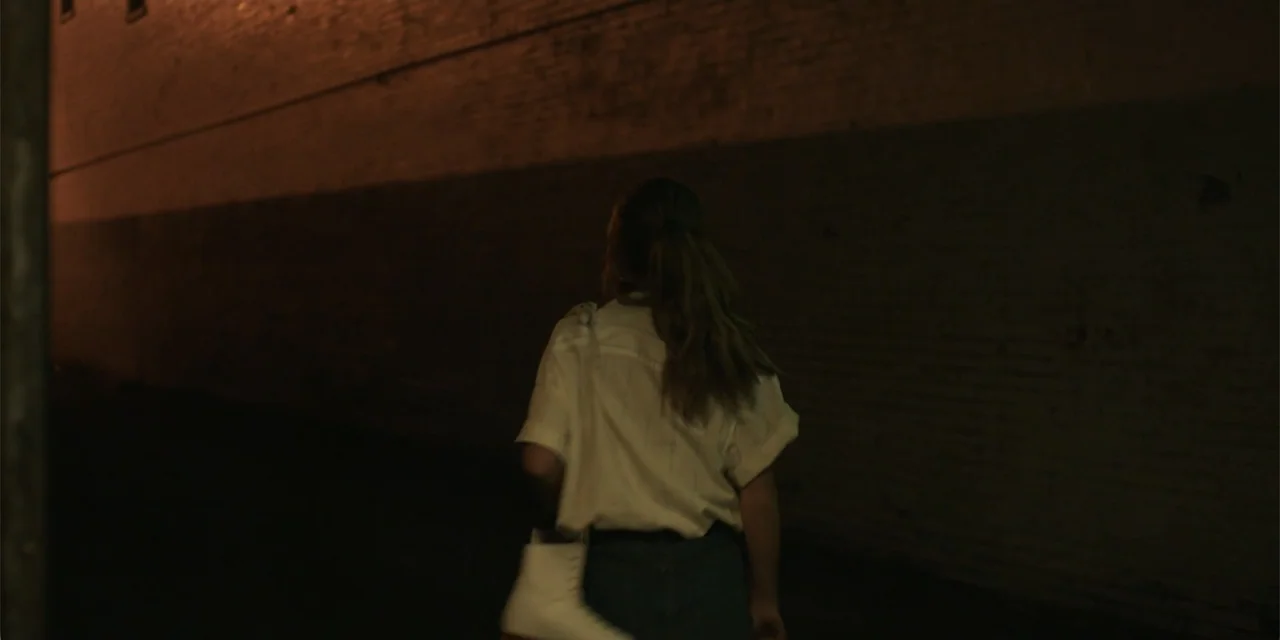 La coda del diavolo (2024) movie still: medium — A dark, narrow alleyway features a long brick wall illuminated by dim light. A young woman walks…
