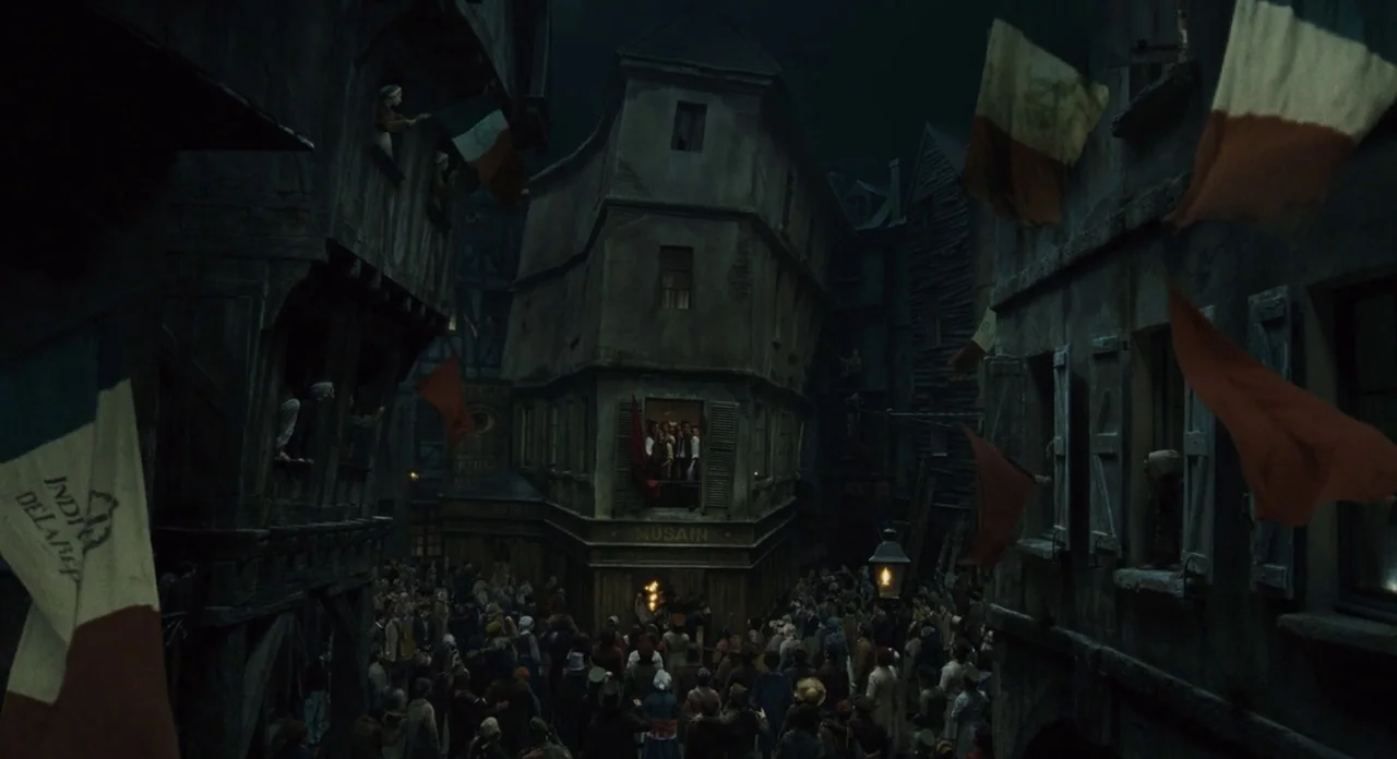 Les Misérables (2012) movie still: establishing — A dense crowd of people occupies a narrow, dark 19th-century street with tall buildings. Red and…