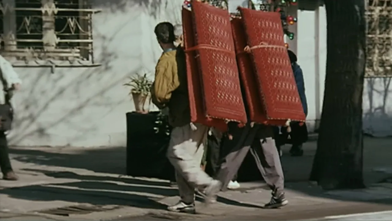 The Mirror (2014) movie still: fullbody — An outdoor urban sidewalk with a building and tree visible in the background. A man walks away from the…