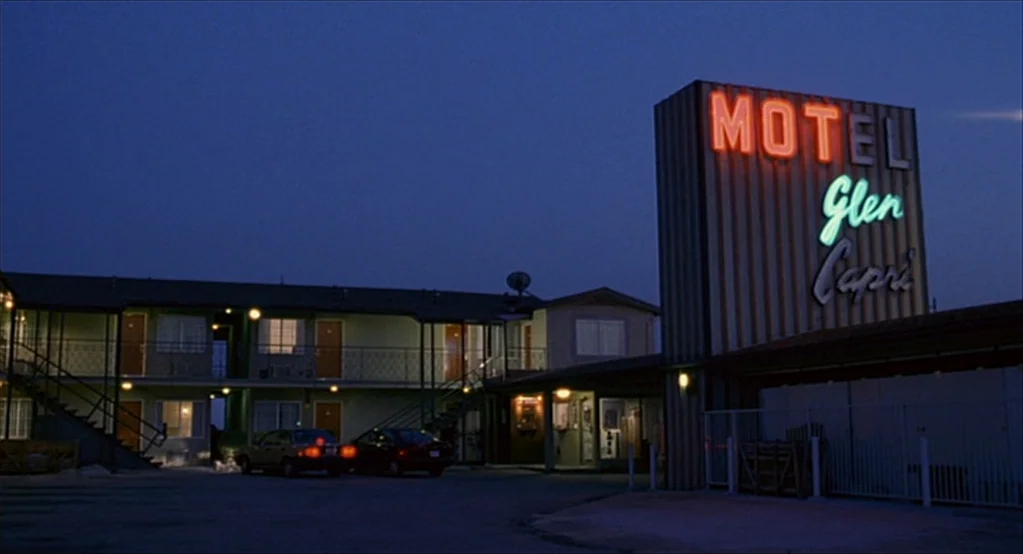 The Good Girl (2002) movie still: establishing — A two-story motel building sits at dusk with a large neon sign attached. Two cars are parked in the…