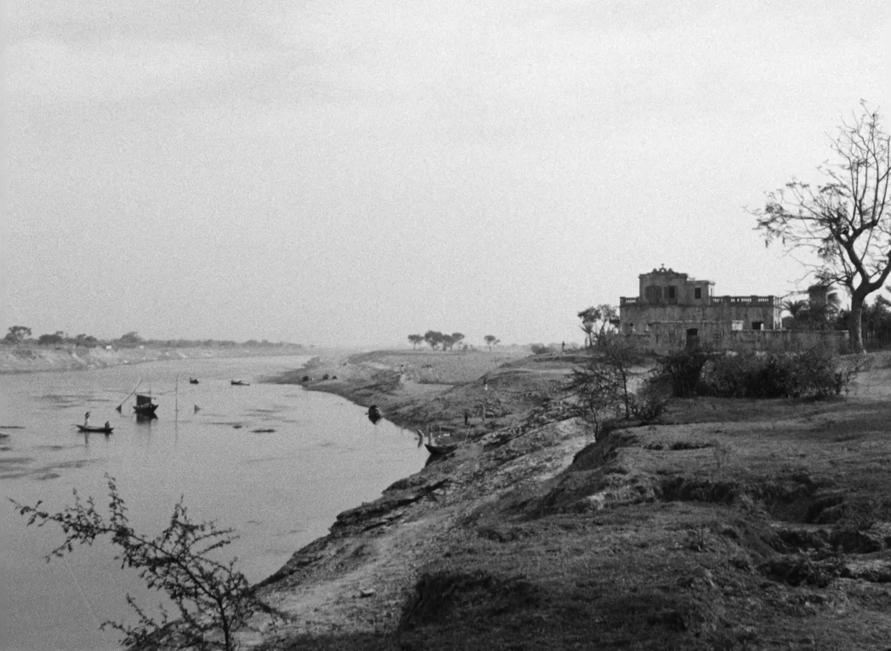 Apur Sansar (1959) movie still: establishing — A wide shot shows a calm river flowing past an arid, grassy embankment. An old, two-story colonial…
