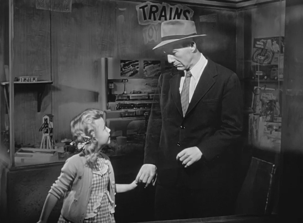 M (1951) (1951) movie still: medium — A dark retail shop with shelves displaying various toy train sets and models. A tall man in a hat and a young…