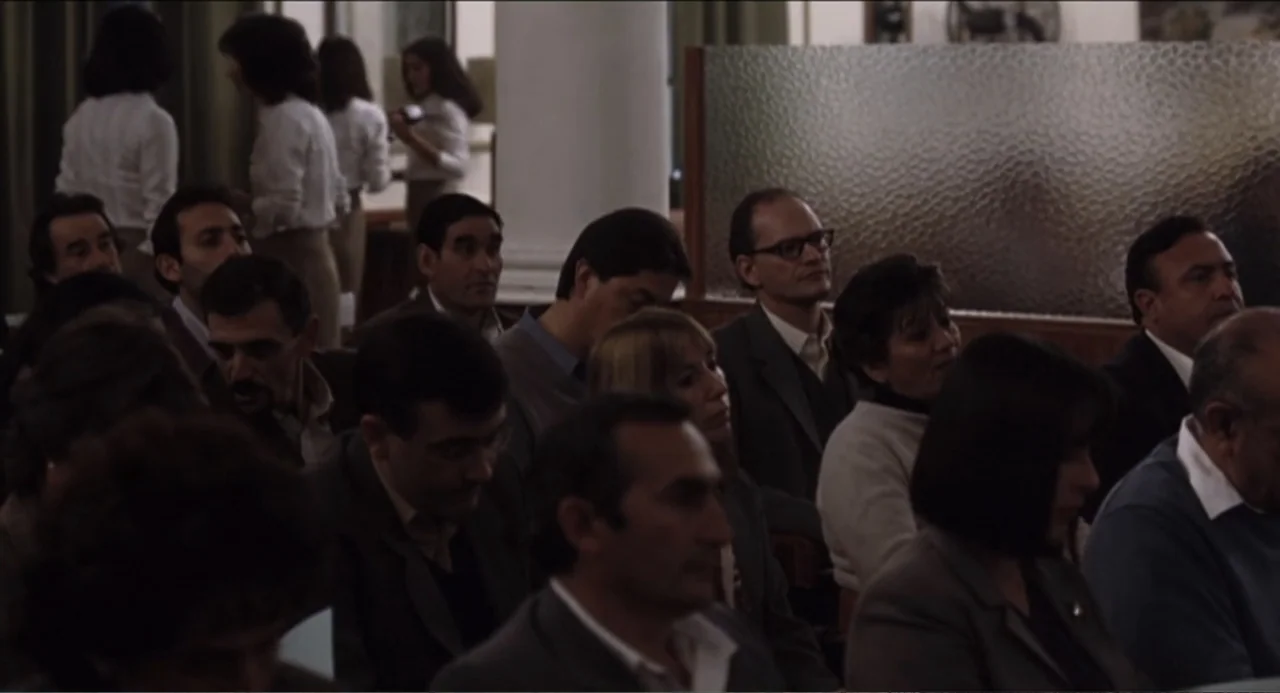 The Holy Girl (2004) movie still: medium — A crowded auditorium interior with rows of people seated under dim indoor lighting. Individuals look…
