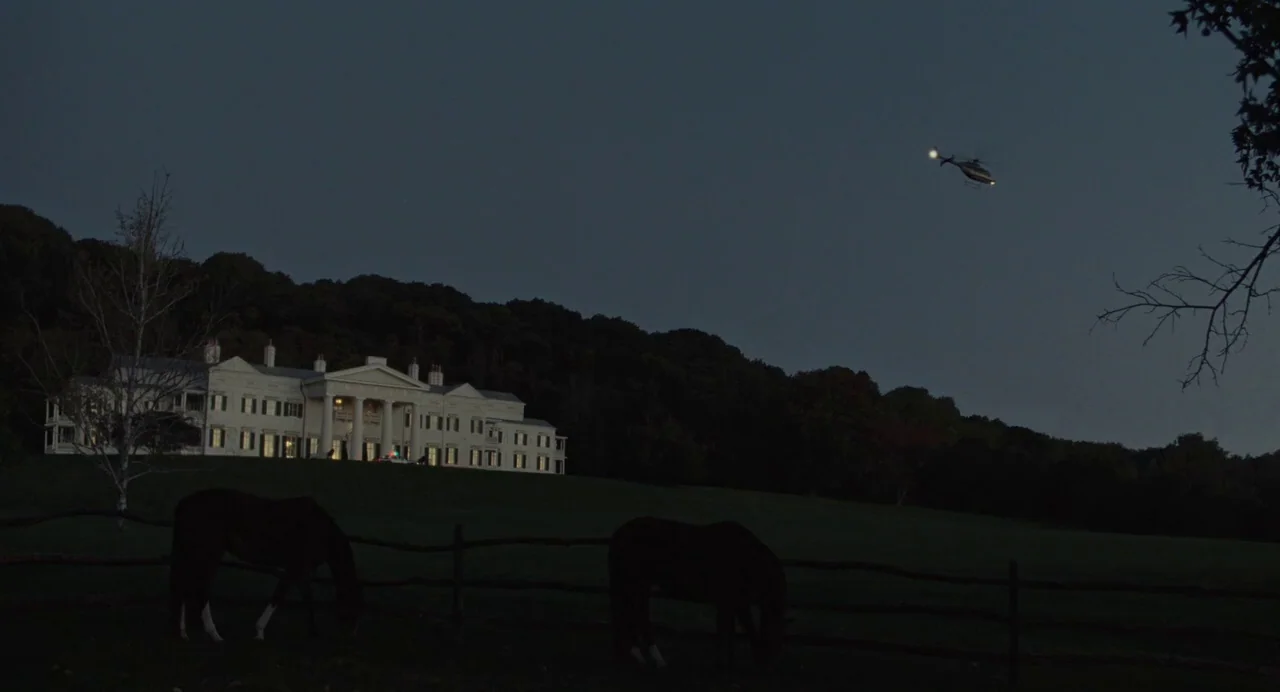 Foxcatcher (2014) movie still: wide — A large white mansion sits on a hill at night, framed by dark trees. A helicopter with an active spotlight…