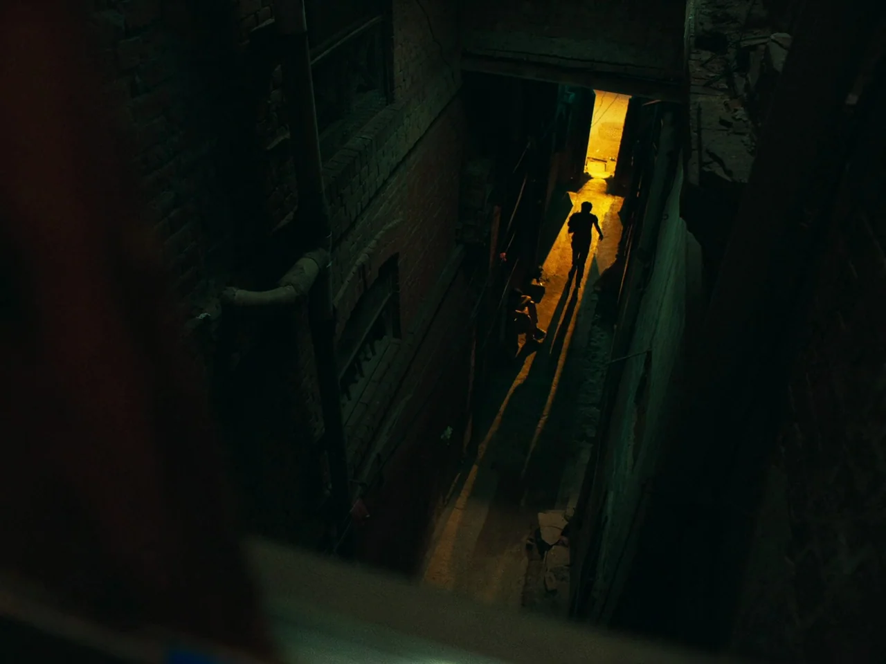 Joyland (2022) movie still: wide — A dark, narrow passageway between two old brick buildings is shadowed and poorly lit. A person walks away from…
