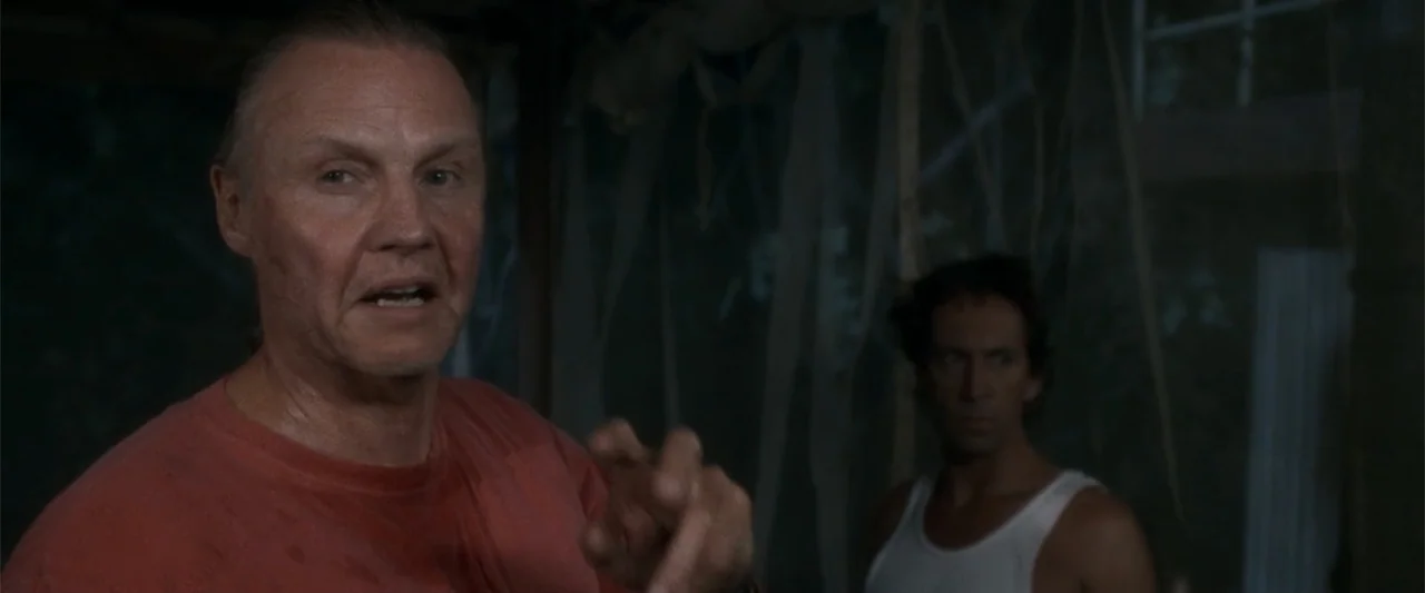 Anaconda (1997) movie still: medium — A dimly lit, rustic structure with weathered vertical wood slats in the background. An older man in a red…
