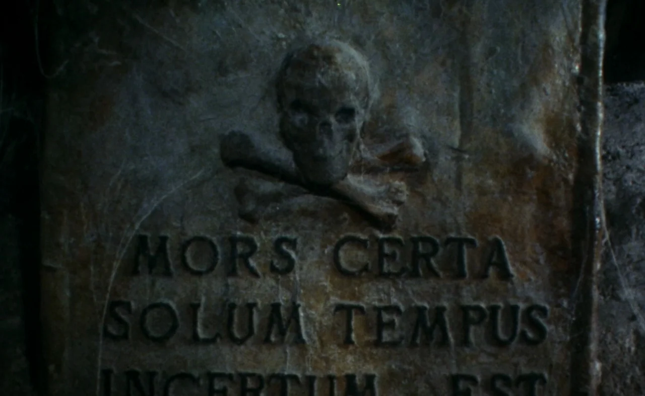 The Pit and The Pendulum (1991) movie still: closeup — A weathered stone grave marker features an engraved skull and crossbones symbol. The latin…