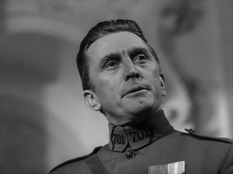 Paths of Glory (1957) movie still: closeup — A man in a military uniform is seen in a dimly lit indoor space. He is looking up and away, appearing…