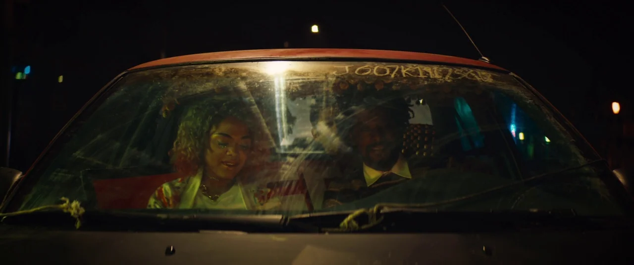 Sorry to Bother You (2018) movie still: medium — A man and woman sit inside the front seat of a dimly lit car. The windshield is smudged and…