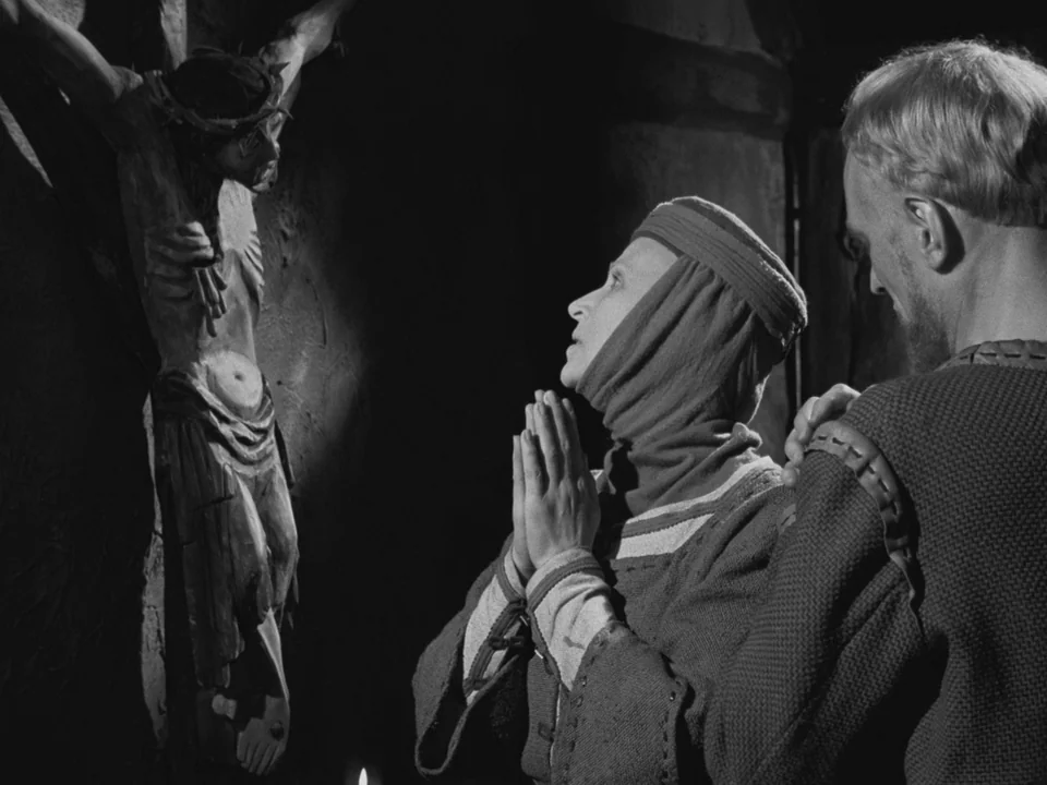 The Virgin Spring (1960) movie still: medium — A dark stone interior features a mounted wooden crucifix illuminated by a single light source. A…