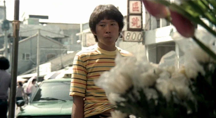The Boys From Fengkuei (1983) movie still: medium — An urban street setting features old signage and out-of-focus flowers in the foreground. A young…