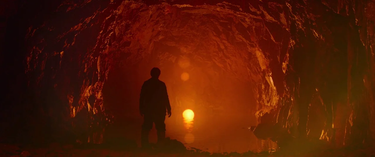Siberia (2019) movie still: wide — An underground cavern has jagged stone walls and a small pool of dark water. A glowing circular light rests on…