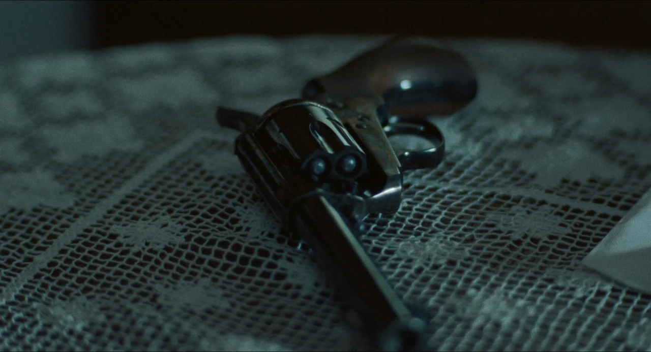 The Lodge (2019) movie still: closeup — A dark, metallic revolver sits atop a white crochet lace doily on a surface. The object is angled slightly…