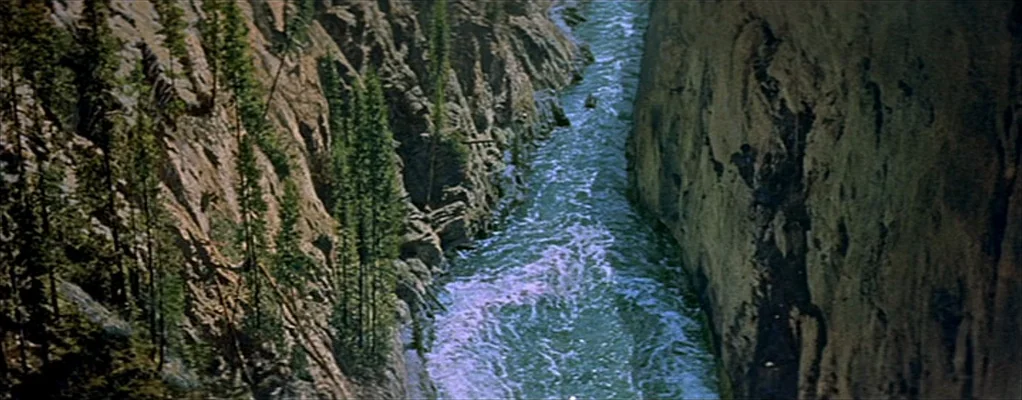 River of No Return (1954) movie still: establishing — A narrow river surges through a deep, steep-walled rocky canyon. Tall evergreen trees grow…