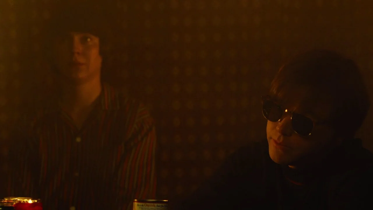 Love & Mercy (2014) movie still: medium — A dimly lit room features dark walls with a subtle pattern and sparse light. Two young men sit near each…