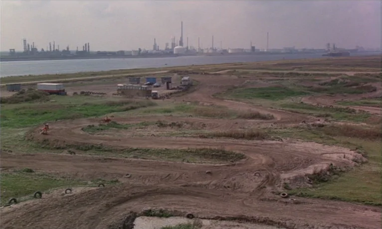 Spetters (1980) movie still: establishing — A wide, outdoor motocross track made of packed earth runs along a large river. An industrial complex…